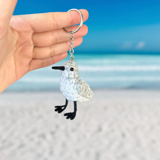 Hand holding a keychain with a white bird design against a beach background