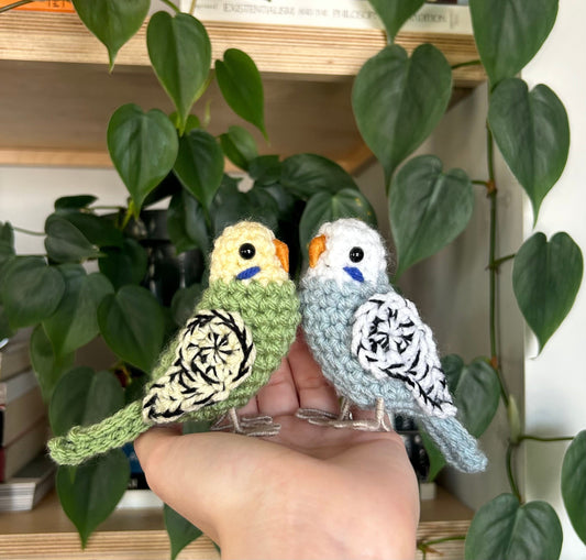 Two handmade parakeet toys held in a hand with green leaves in the background