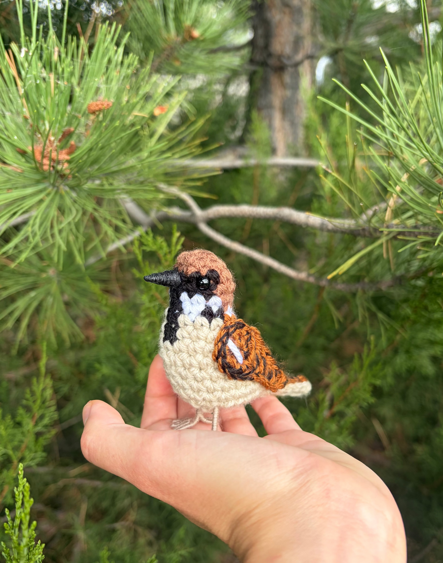 A small crocheted bird figurine perched on a person's hand, with a blurred forest background of pine branches and green needles.