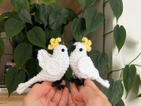 Two white crochet birds with yellow accents held between two hands against a green leafy background.