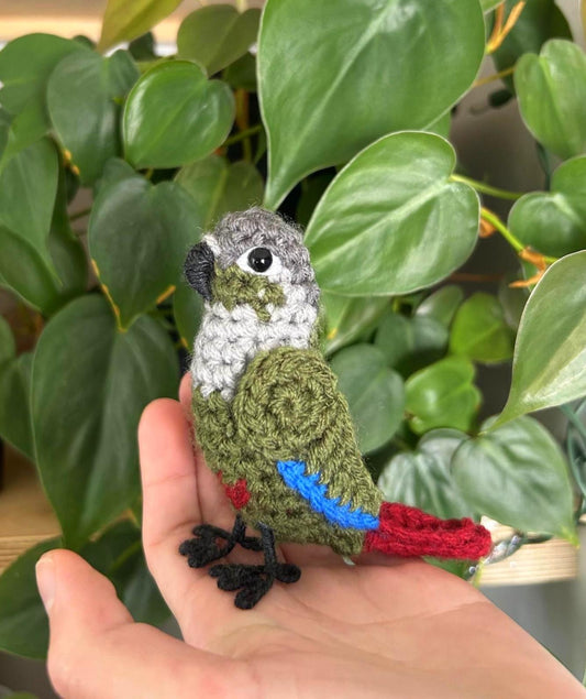 Hand holding a small crocheted parrot toy in front of green leaves