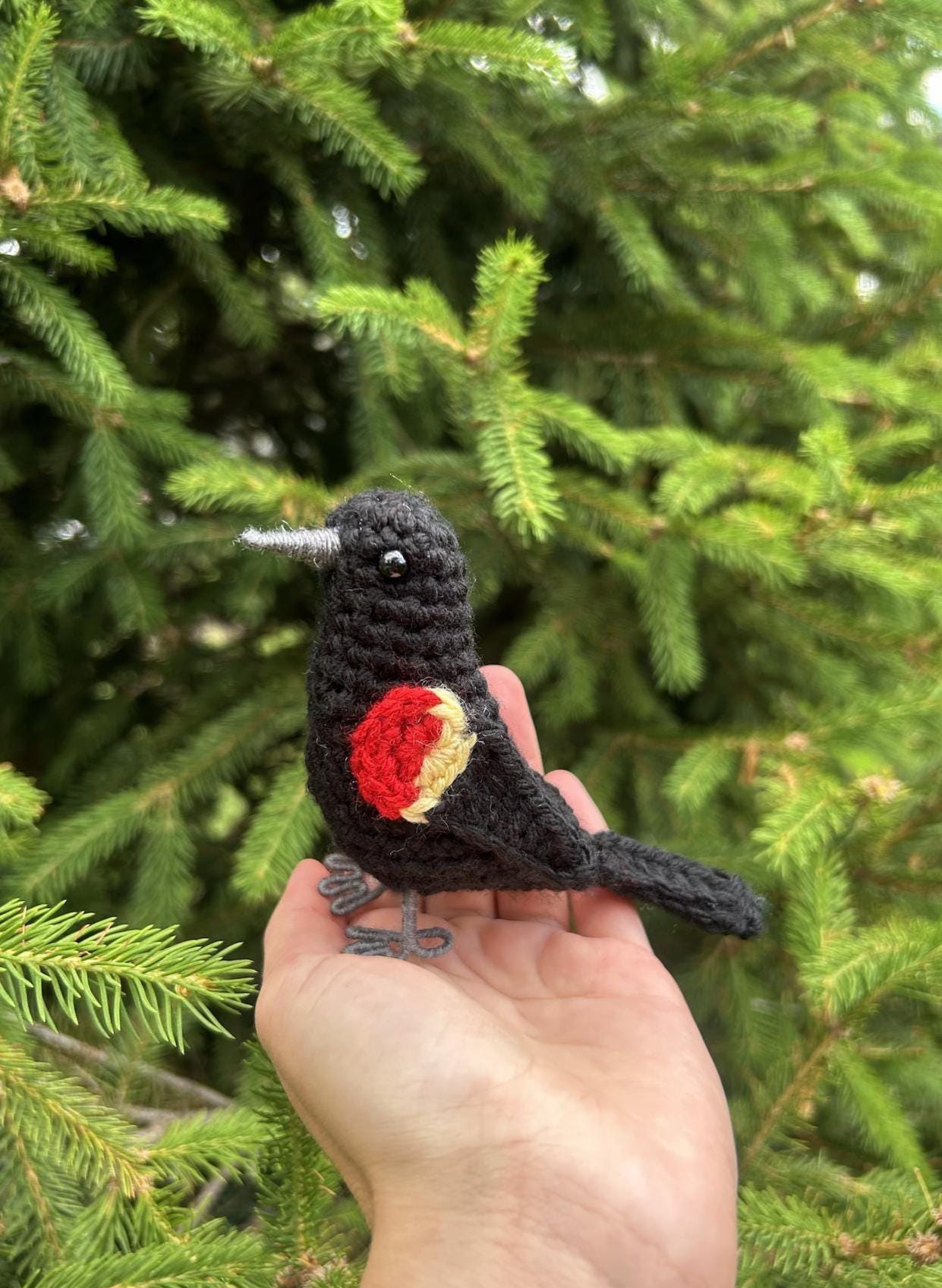 Hand holding a black crochet bird with red and yellow details against a green tree background