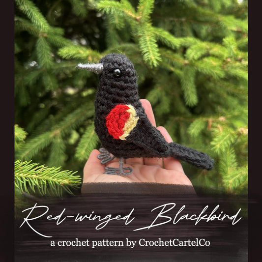 A crochet red-winged blackbird crochet pattern shown in front of green foliage, with 'CrochetCartelCo' branding.