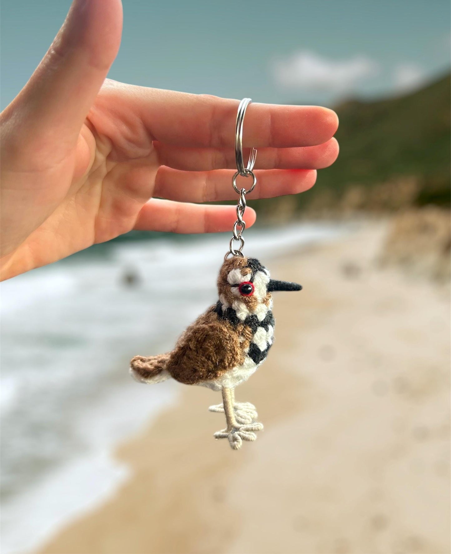 Hand holding a keychain with a bird design on a beach.