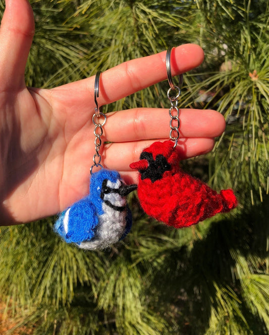 Hand holding two crochet bird keychains, one blue and one red, against a green foliage background.
