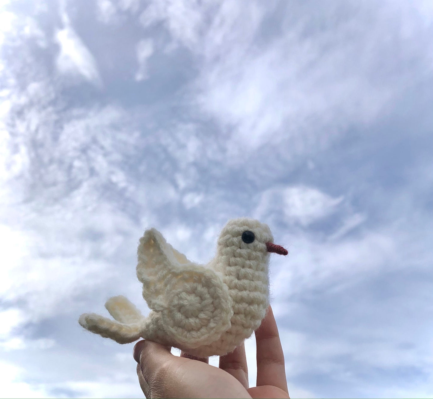 Hand holding a small crochet dove against a cloudy sky