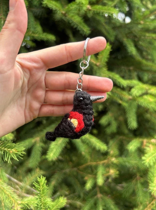 Hand holding a black and red crochet bird keychain against a green tree background