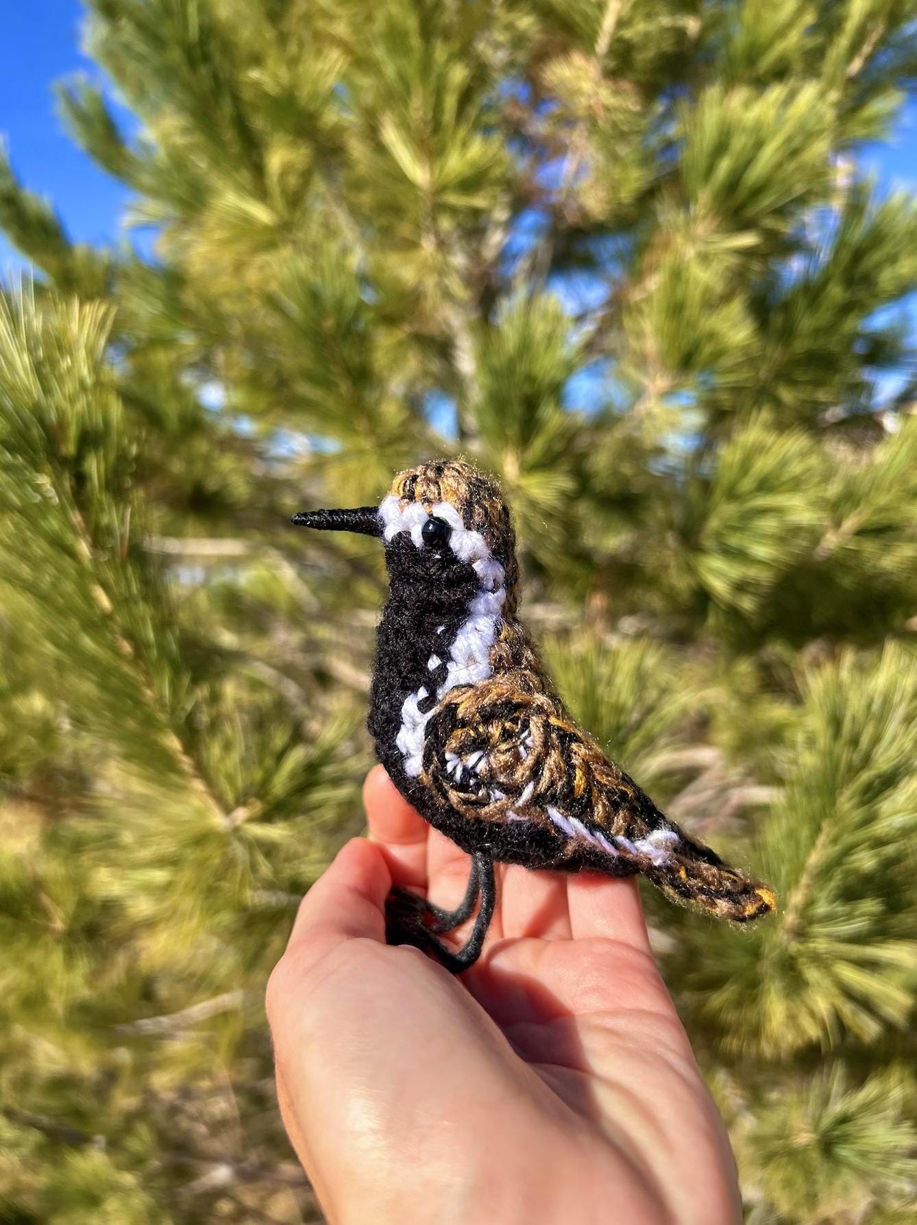 Hand holding a crocheted bird in front of a pine tree