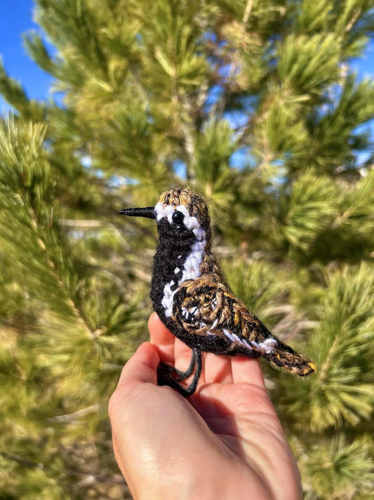 Hand holding a crocheted bird in front of a pine tree
