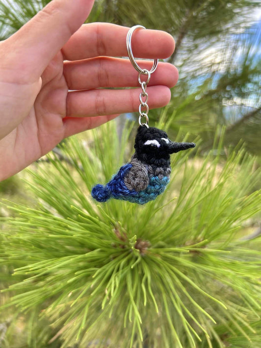 Hand holding a crochet bird keychain against a green pine tree background