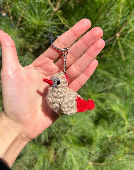 Hand holding a small bird-shaped keychain with red accents against a green leafy background