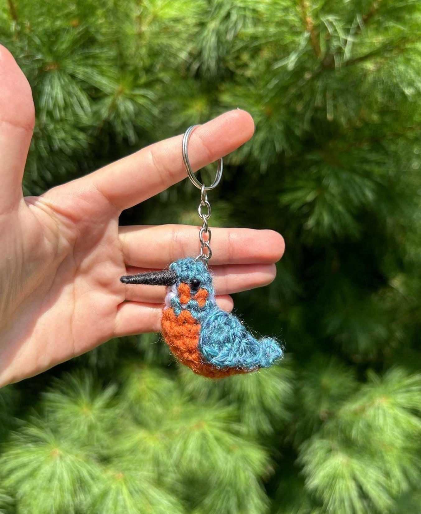 Hand holding a blue and orange bird-shaped keychain against a green tree background