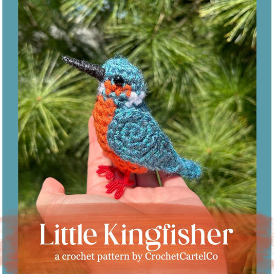 Crocheted kingfisher bird held in a hand with a green leafy background, featuring 'Little Kingfisher' pattern by CrochetCartelCo.