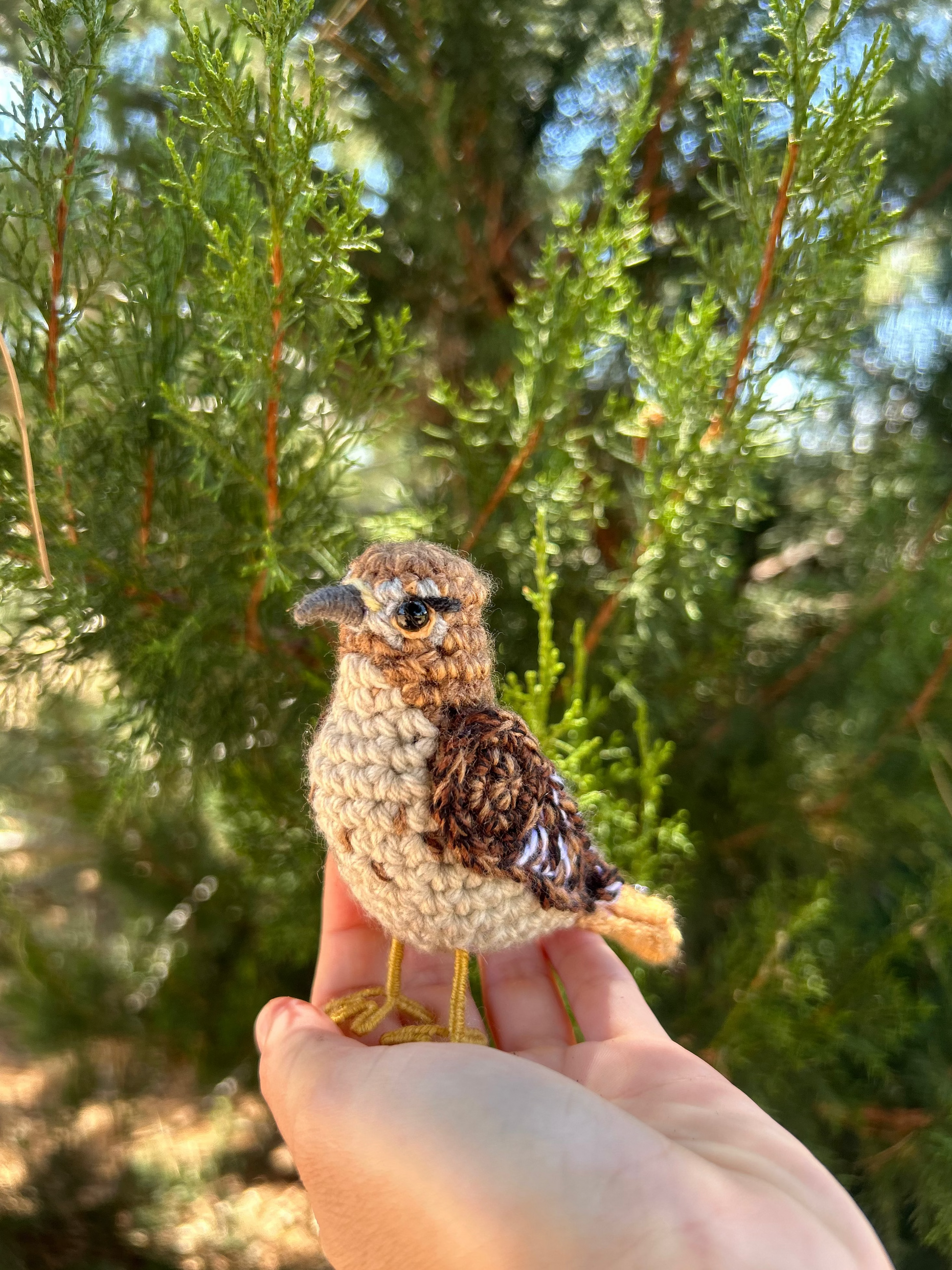A small crocheted bird figurine with brown and beige feathers is being held on a person's hand outdoors in front of green foliage.