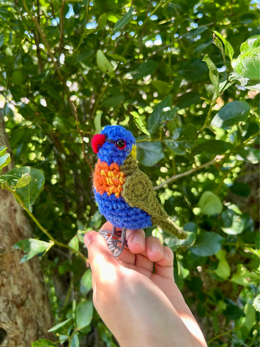 A small crocheted bird figurine in bright blue, orange, red, and olive green is being held in a person's hand outdoors in front of leafy green trees.