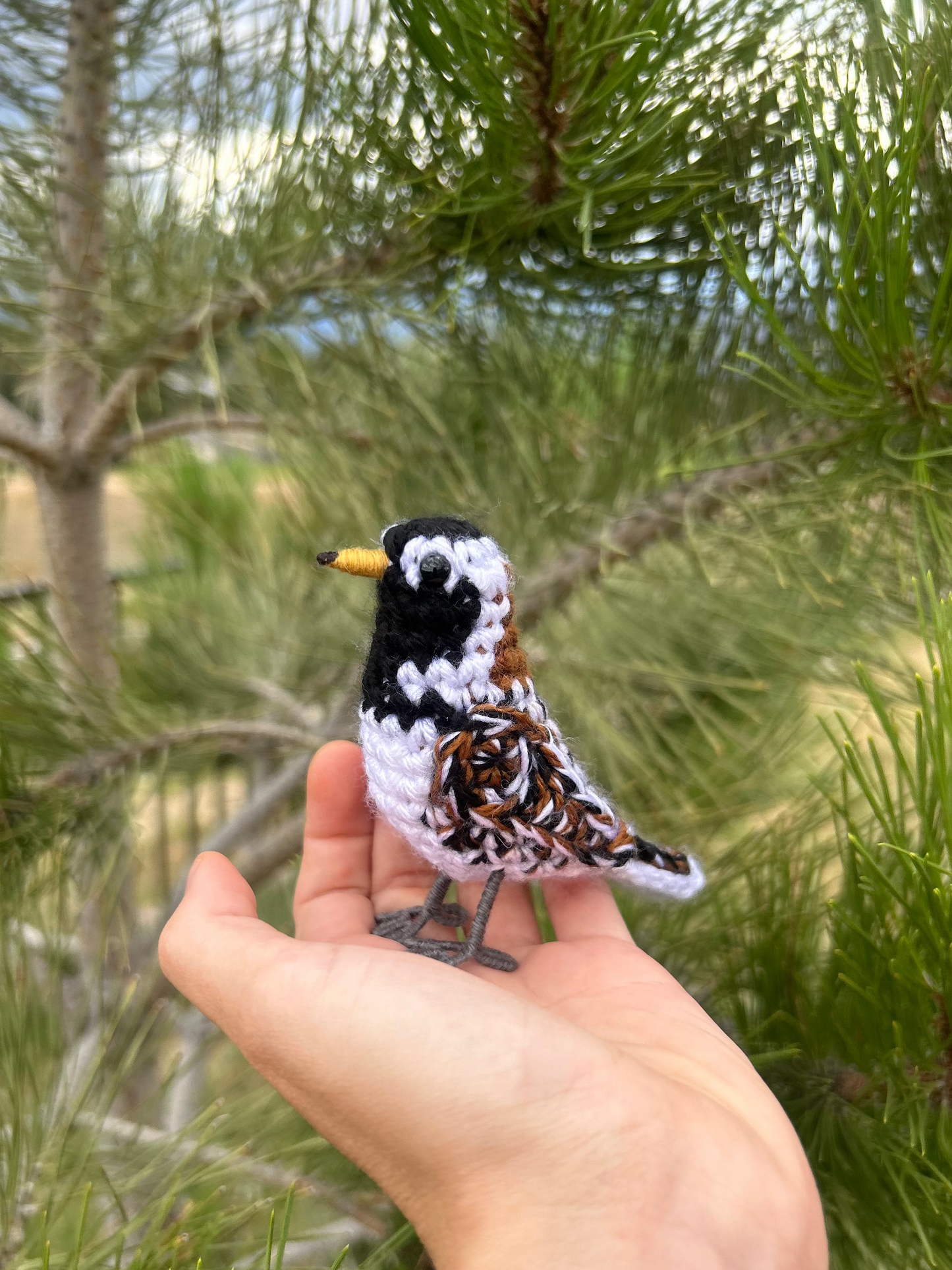 A small handmade crochet bird with a black, white, and brown patterned body and a yellow beak, resting on a person's hand with pine branches in the background.
