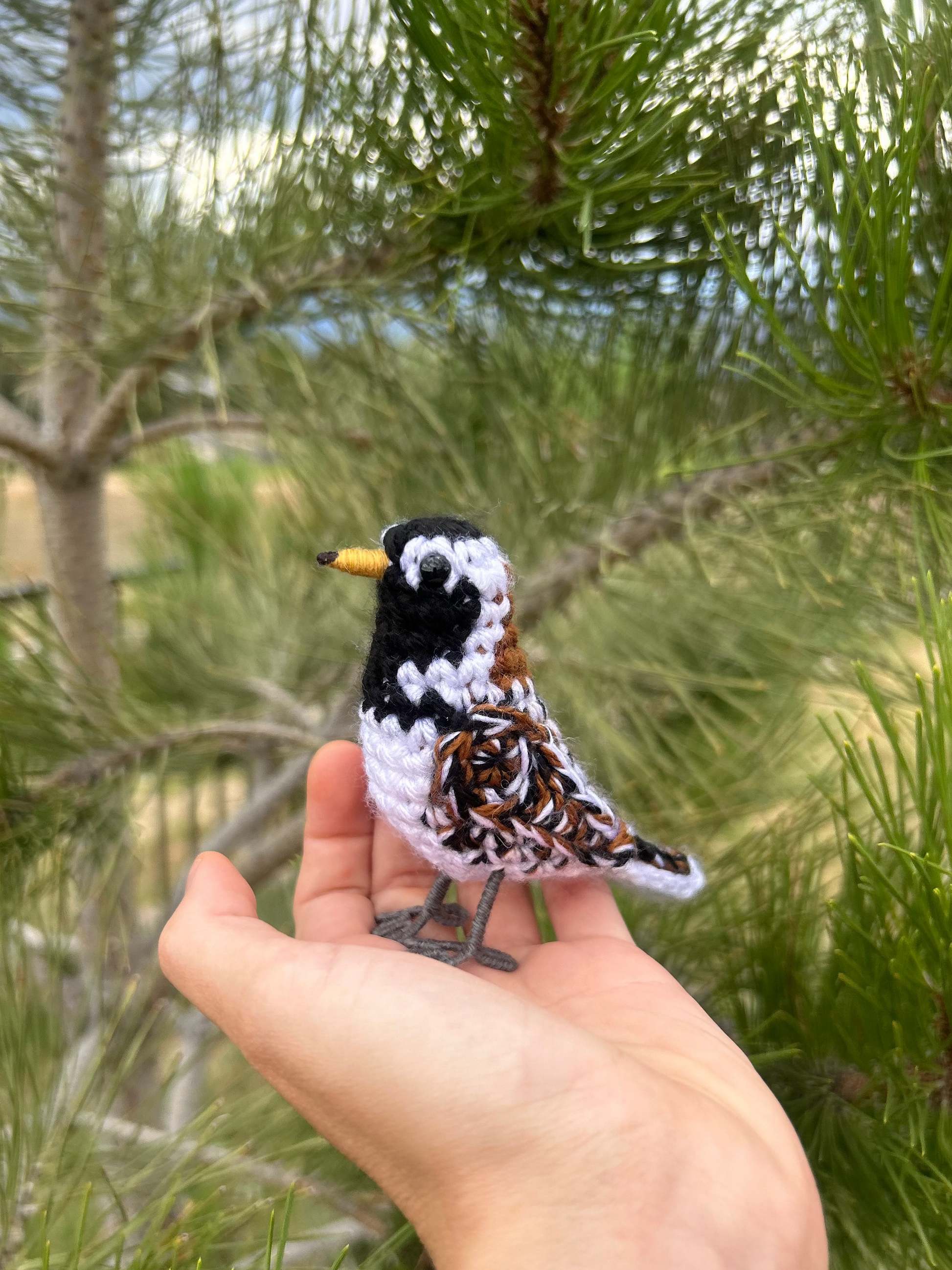 A small handmade crochet bird with a black, white, and brown patterned body and a yellow beak, resting on a person's hand with pine branches in the background.