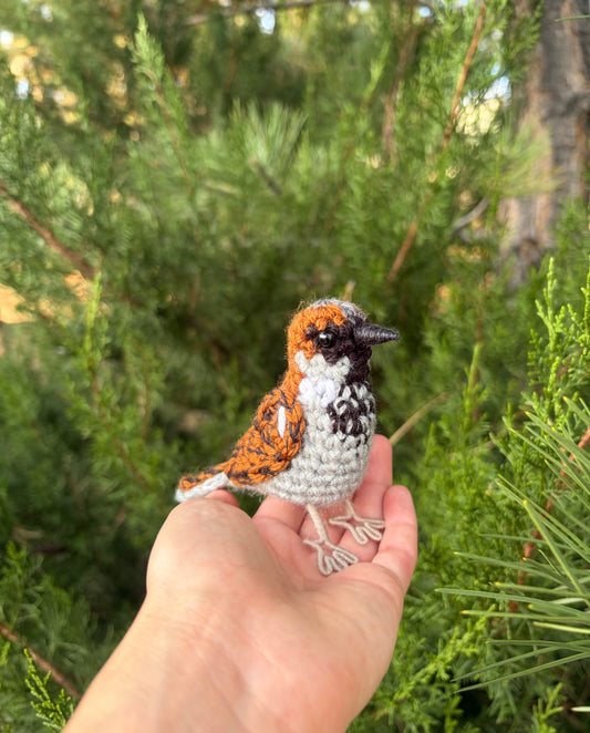 A small crocheted house sparrow bird figurine is held in a person's hand against a background of green pine foliage.