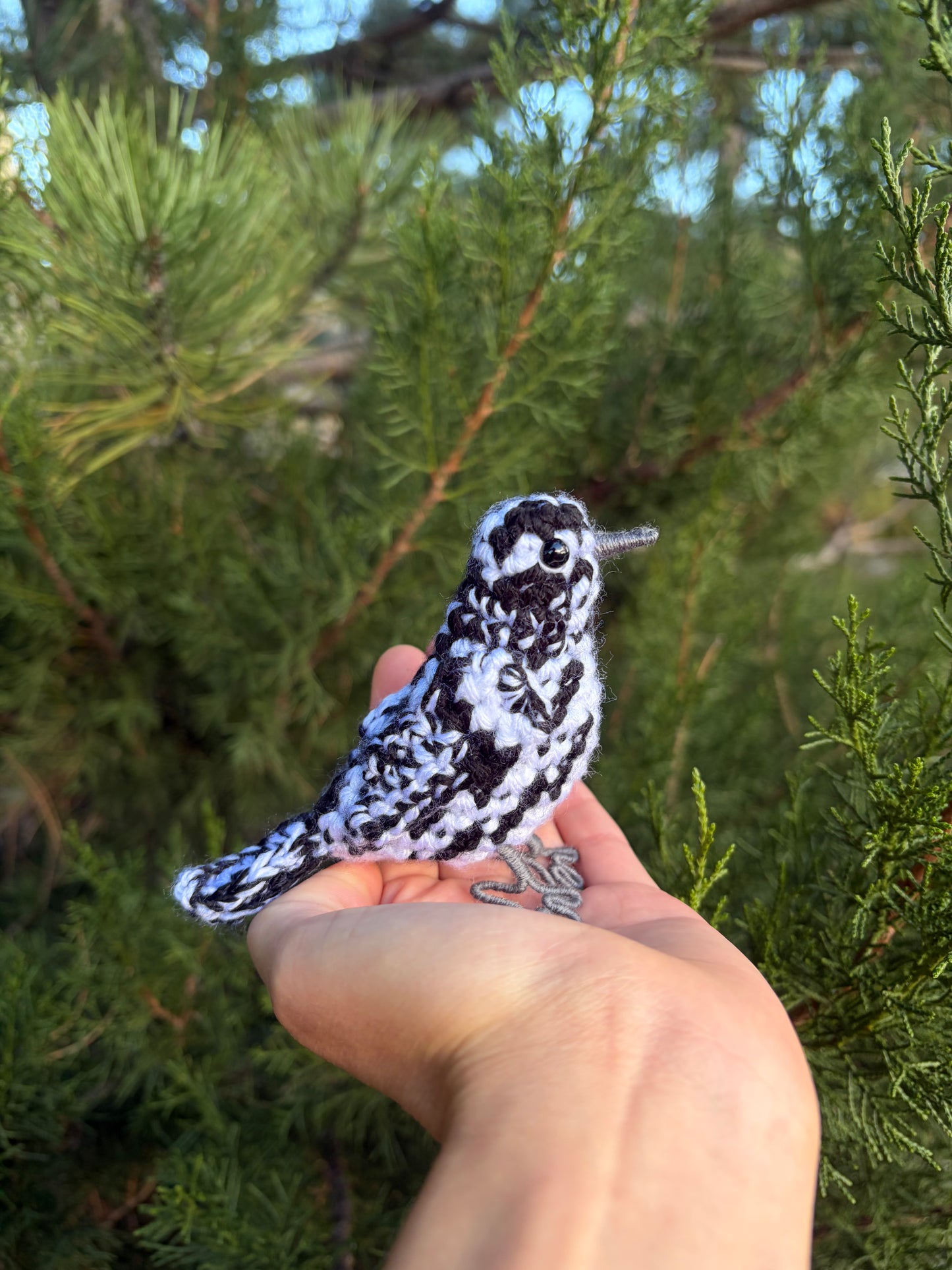 A small black-and-white patterned bird figurine is being held in a person's hand outdoors in front of green pine branches.
