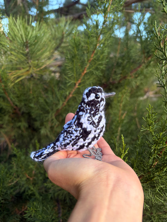 A small black-and-white patterned bird figurine is being held in a person's hand outdoors in front of green pine branches.