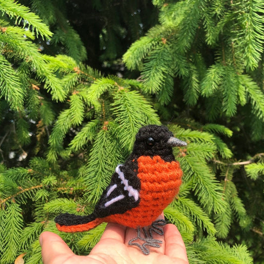 Hand holding a small crocheted bird against a green tree background