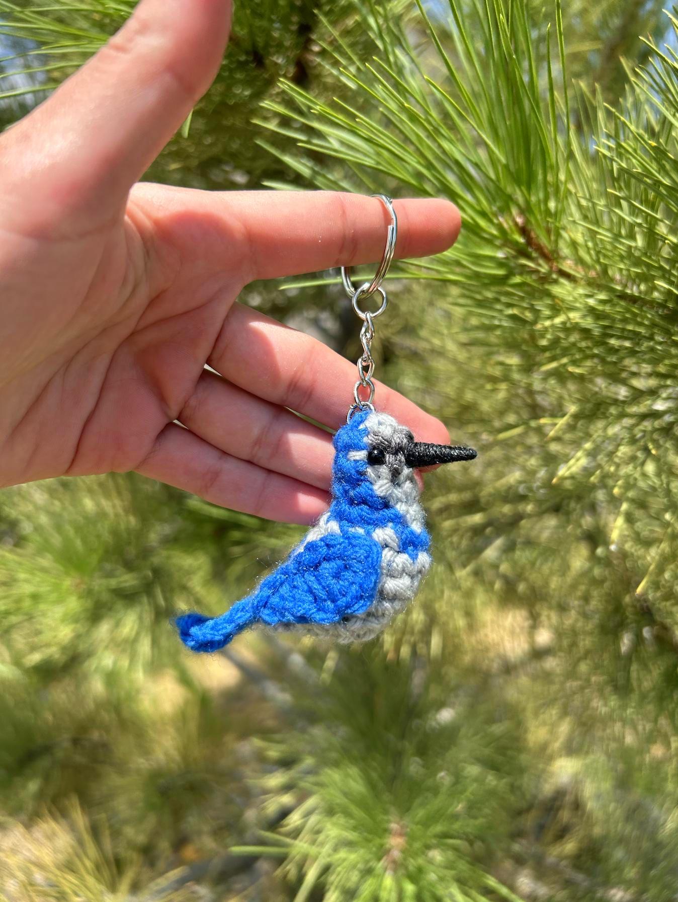 Hand holding a blue and gray bird keychain against a green pine tree background