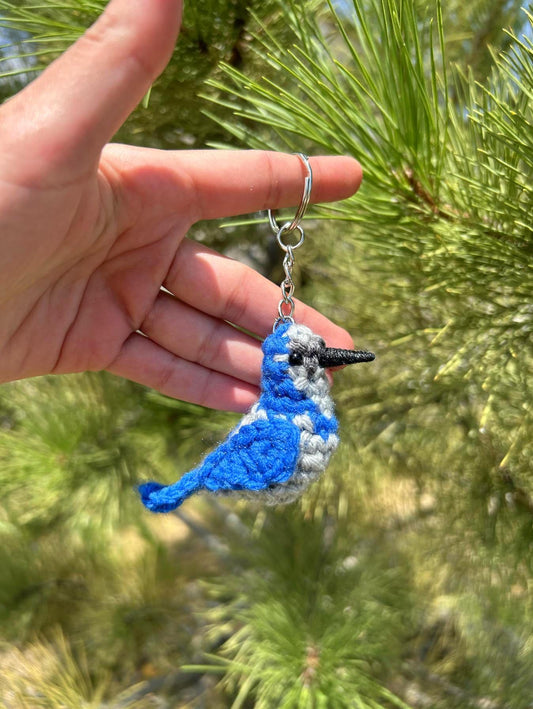 Hand holding a blue and gray bird keychain against a green pine tree background