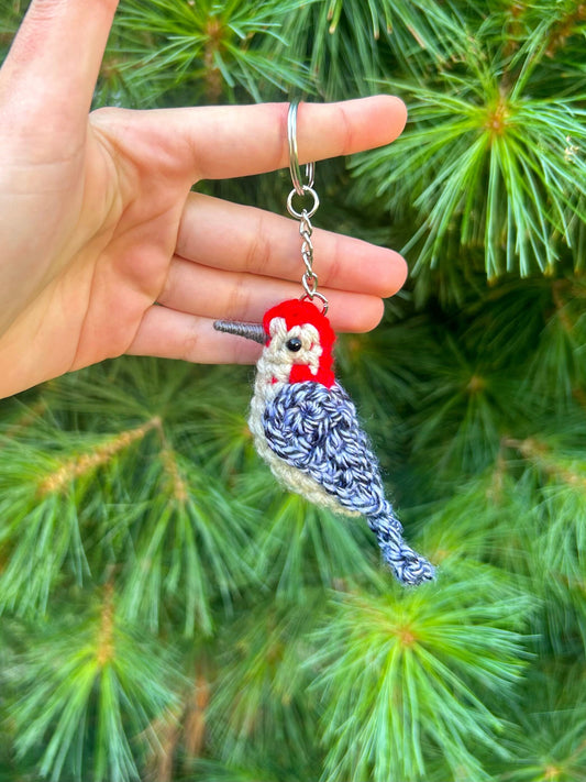 Hand holding a crochet woodpecker keychain against a green pine tree background