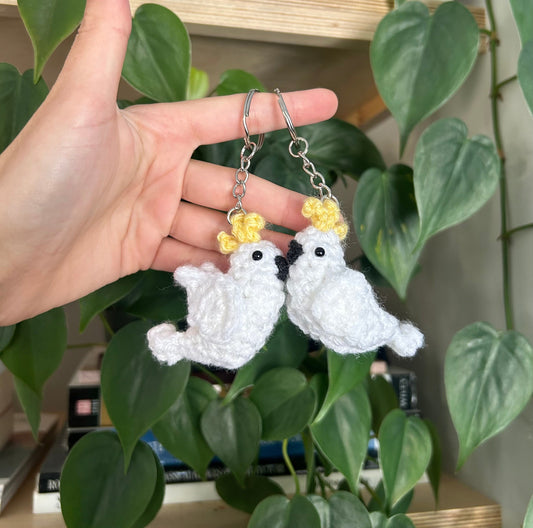 Hand holding two cockatoo keychains against a green leafy background.