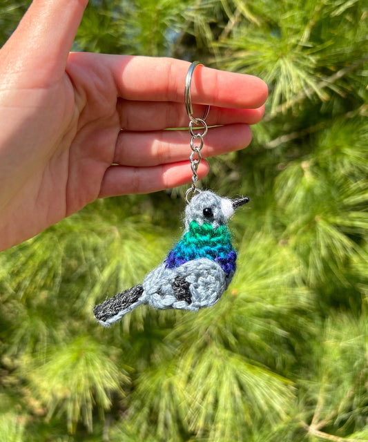 Hand holding a crocheted bird keychain with a green and blue color pattern against a blurred natural background.