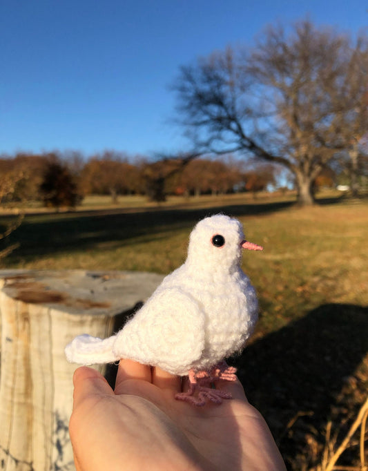 Crocheted white pigeon held in a hand with a scenic background of trees.